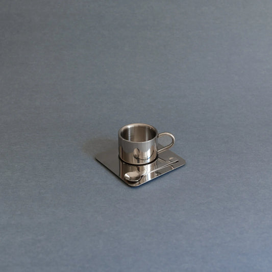 A small metallic silver coffee cup-shaped object resting on a square matching base with a spoon detail, photographed on a plain gray background.