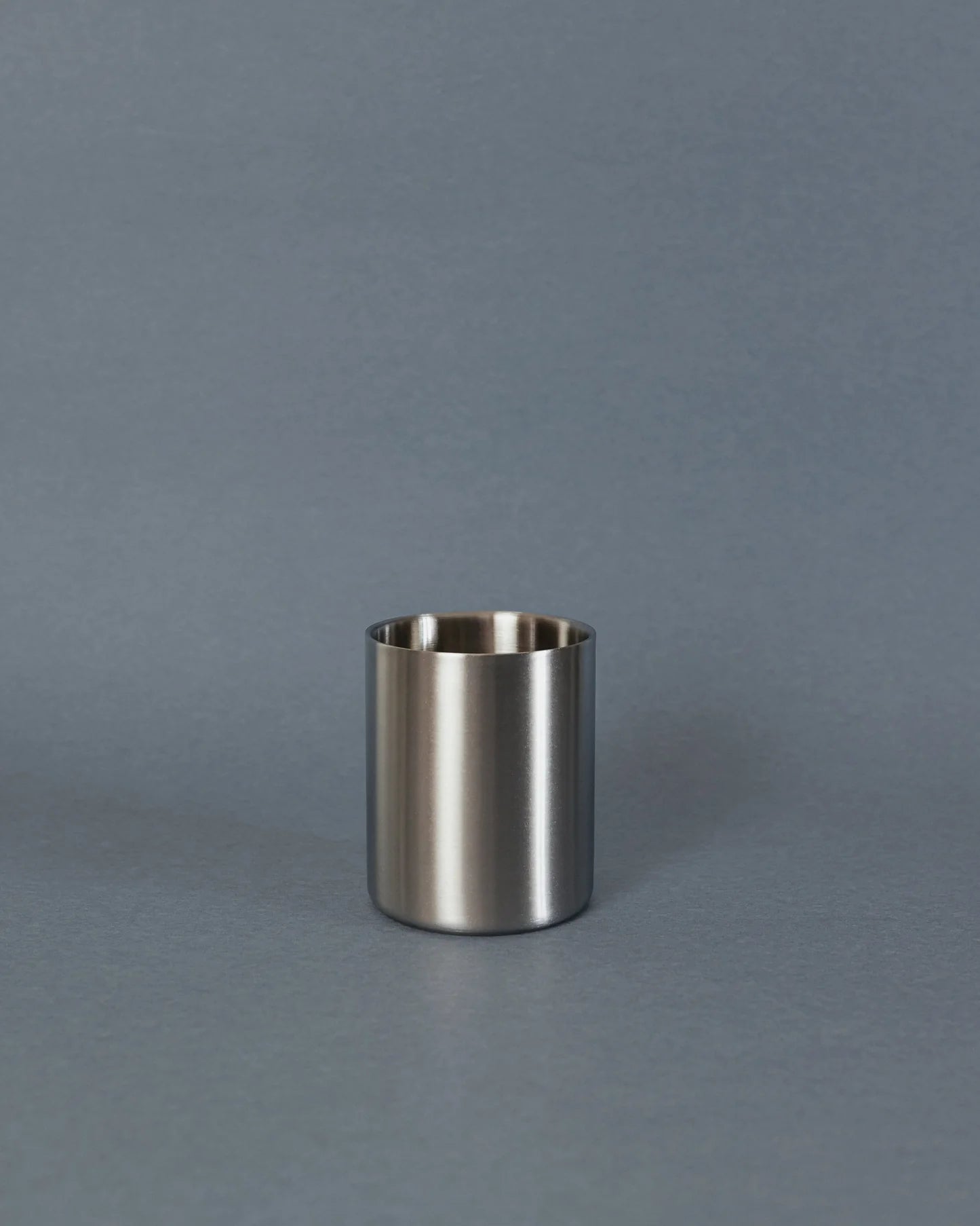 A small brushed metal cup or tumbler with a matte silver finish, standing upright on a smooth gray surface against a matching gray background.
