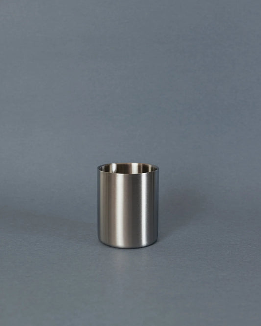 A small brushed metal cup or tumbler with a matte silver finish, standing upright on a smooth gray surface against a matching gray background.