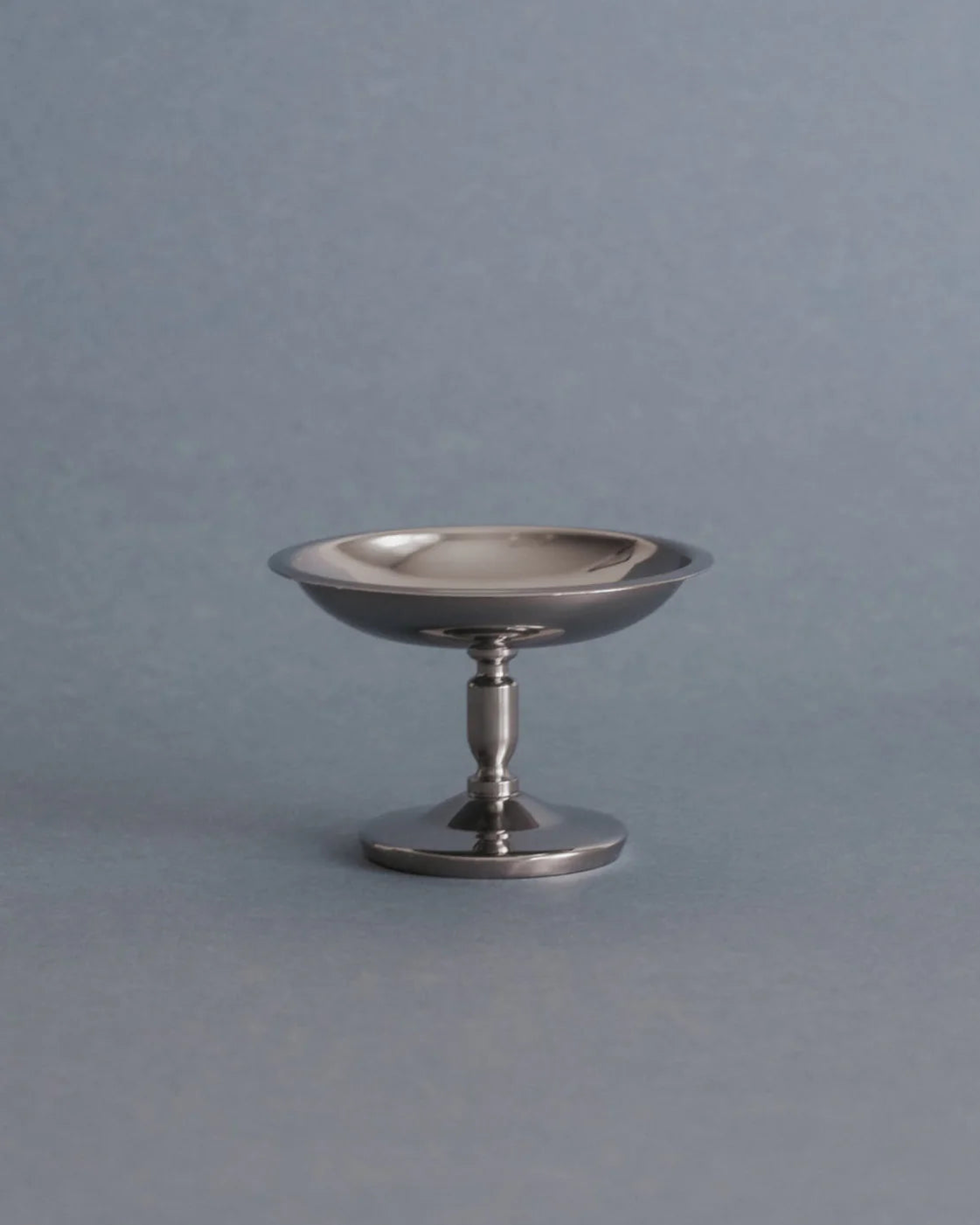 A small polished silver pedestal dish or candle holder with a flared rim, standing on a round base against a smooth gray background.