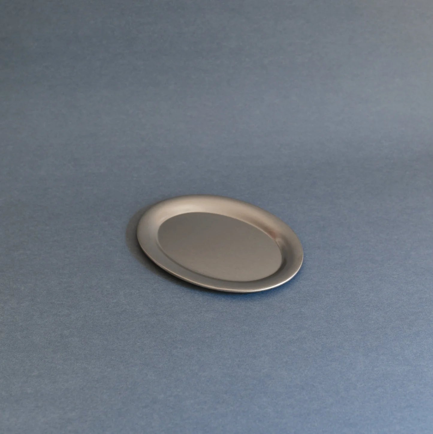 A small oval metallic tray or dish with a brushed silver finish, shown on a smooth slate-blue background.