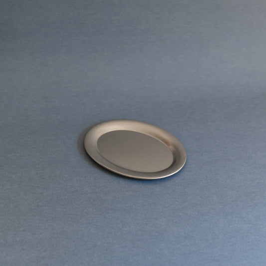 A small oval metallic tray or dish with a brushed silver finish, shown on a smooth slate-blue background.