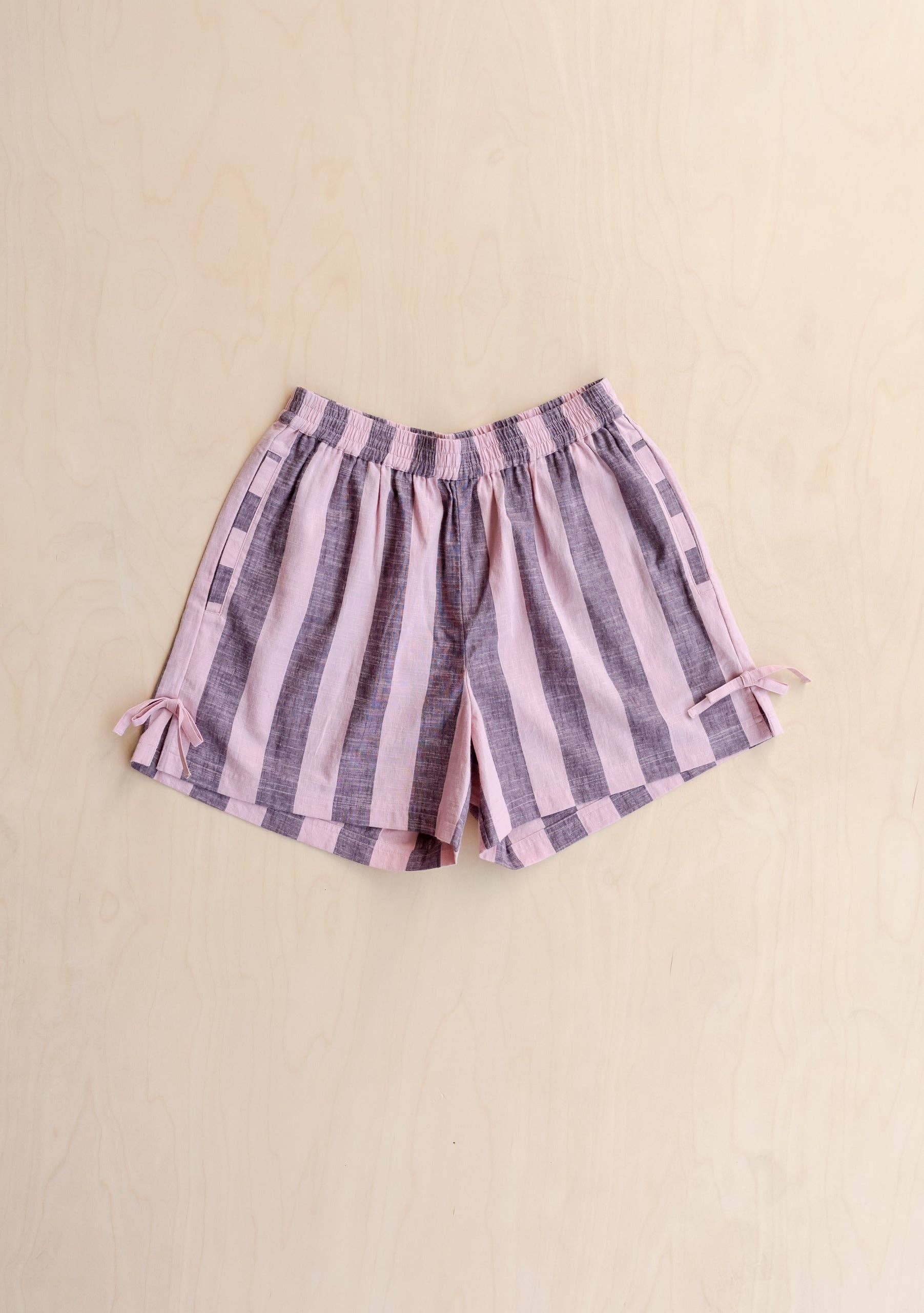 A pair of women's shorts with vertical pink and purple stripes, featuring an elastic waistband and small side ties, laid flat on a light beige background.