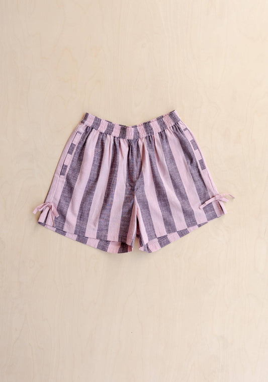 A pair of women's shorts with vertical pink and purple stripes, featuring an elastic waistband and small side ties, laid flat on a light beige background.