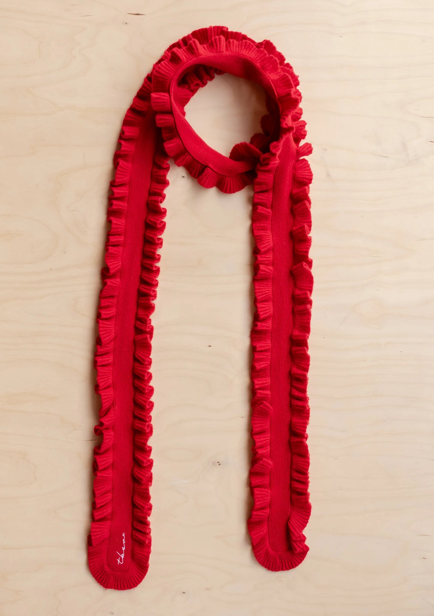 A bright red knit scarf with a continuous ruffled border, styled in a loop on a light-colored wood background.