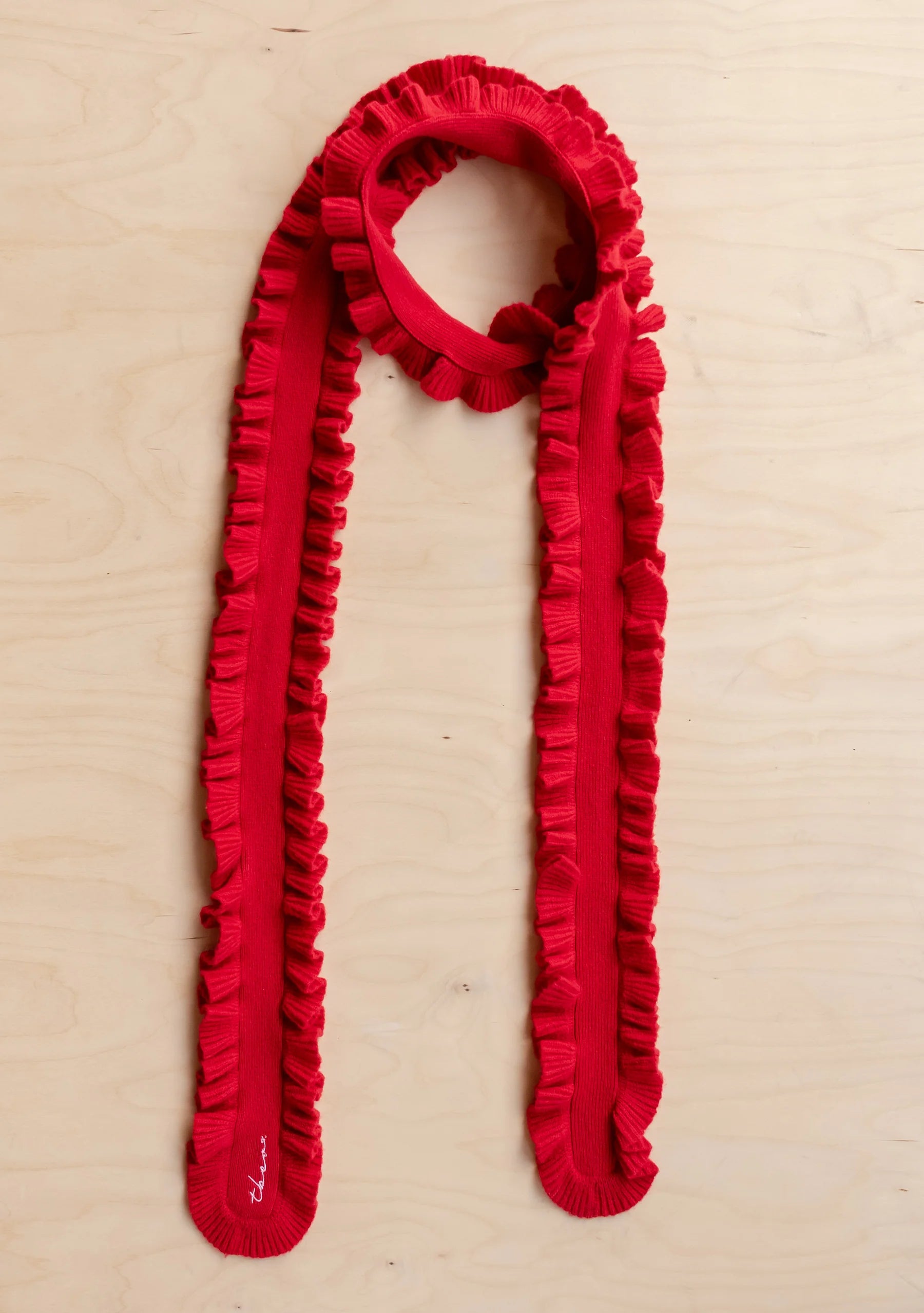 A bright red knit scarf with a continuous ruffled border, styled in a loop on a light-colored wood background.