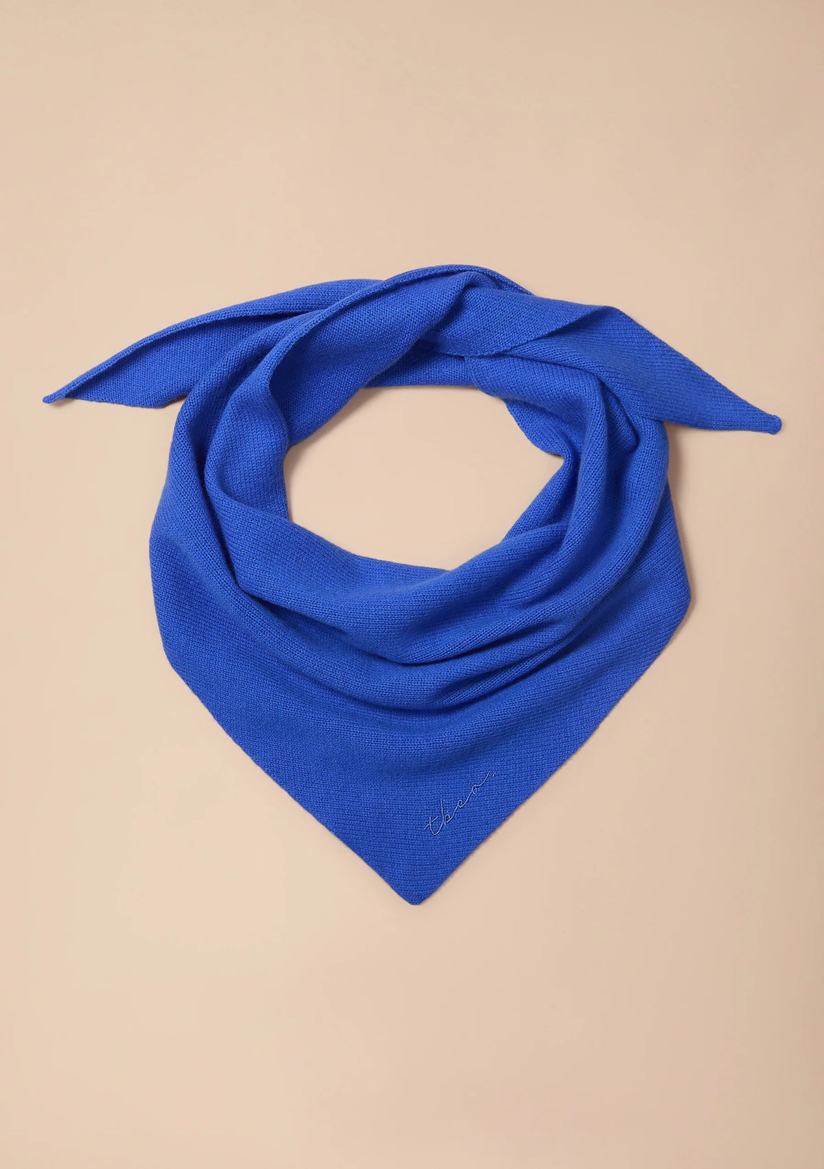 A bright royal blue knit bandana scarf, styled in a loose knot, displayed on a solid beige background.