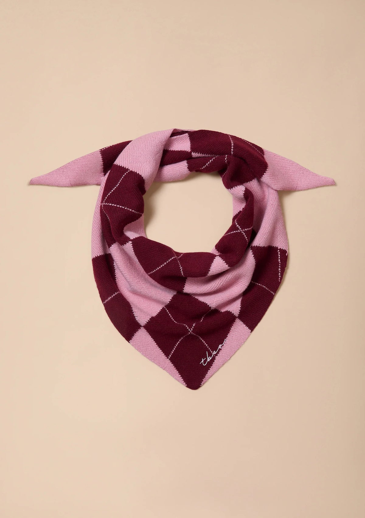 A pink and burgundy argyle knit bandana with white line details, styled in a loop on a solid beige background.
