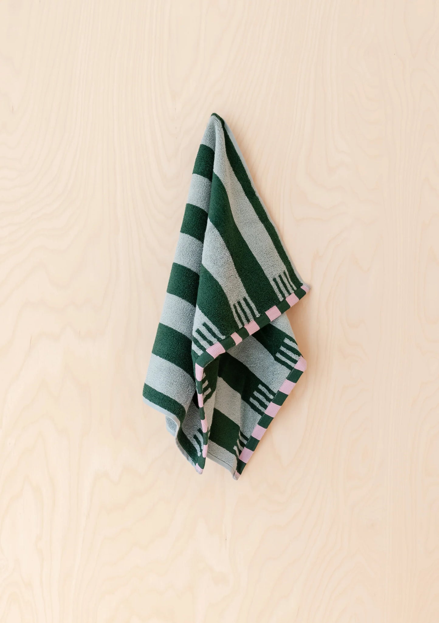 A folded striped textile, likely a small towel or scarf, hanging against a light wood wall. The fabric features alternating dark green and light gray stripes with small pink geometric accents along the edge.