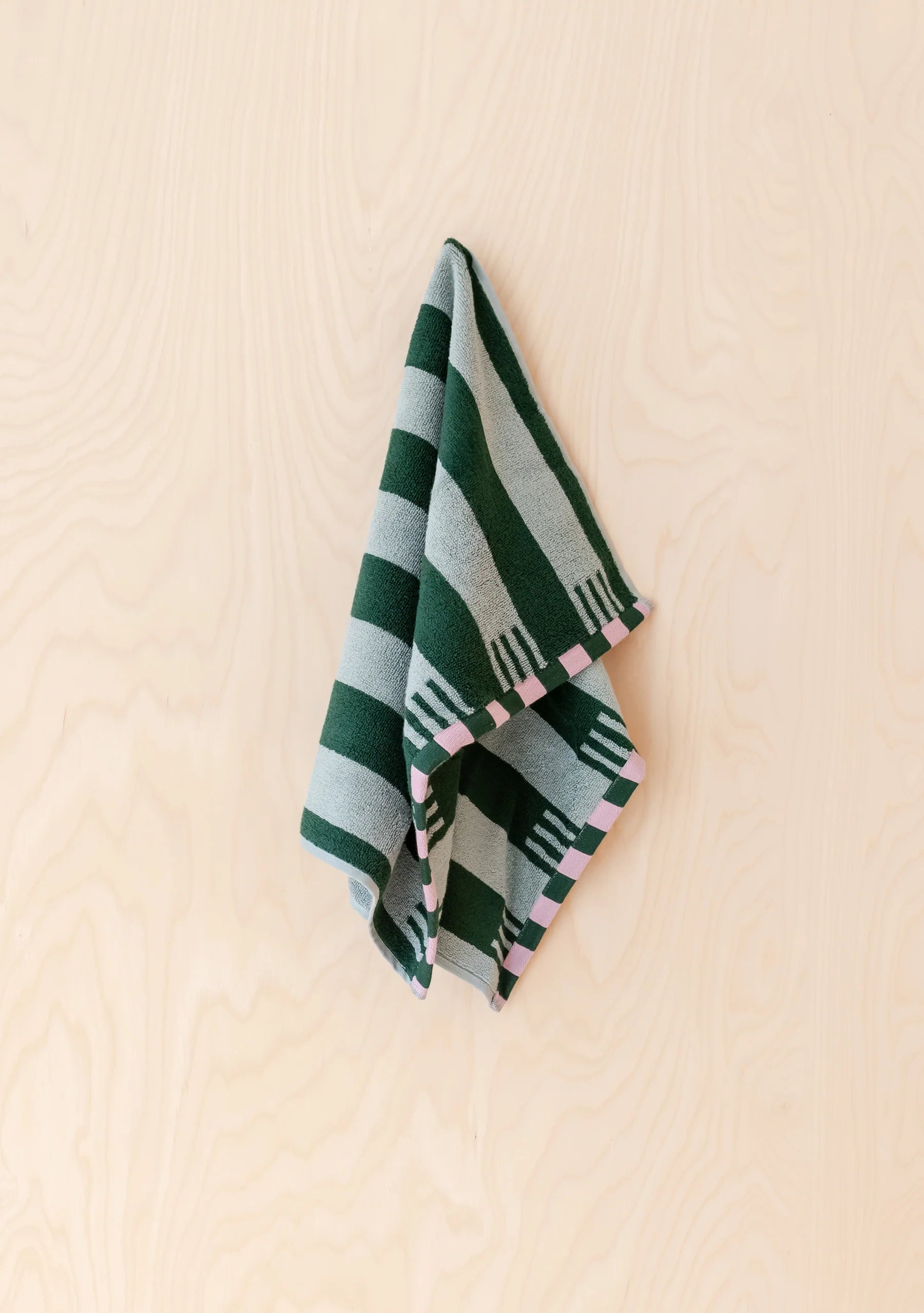 A folded striped textile, likely a small towel or scarf, hanging against a light wood wall. The fabric features alternating dark green and light gray stripes with small pink geometric accents along the edge.