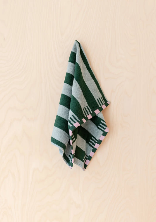 A folded striped textile, likely a small towel or scarf, hanging against a light wood wall. The fabric features alternating dark green and light gray stripes with small pink geometric accents along the edge.