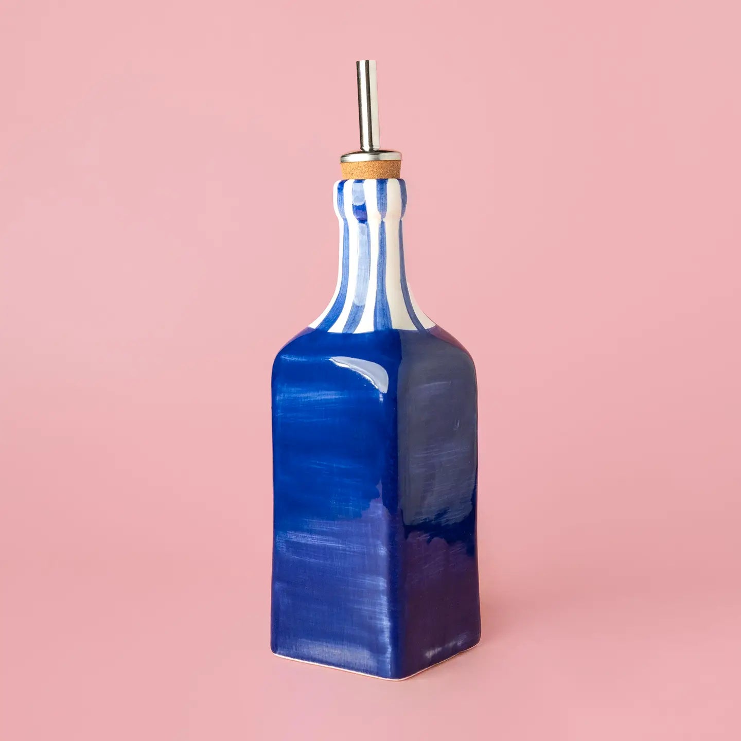 A glossy cobalt blue glass bottle with a flared neck, featuring a white and blue striped pattern on the upper neck and a silver metal pour spout with a gold collar, set against a solid pink background.