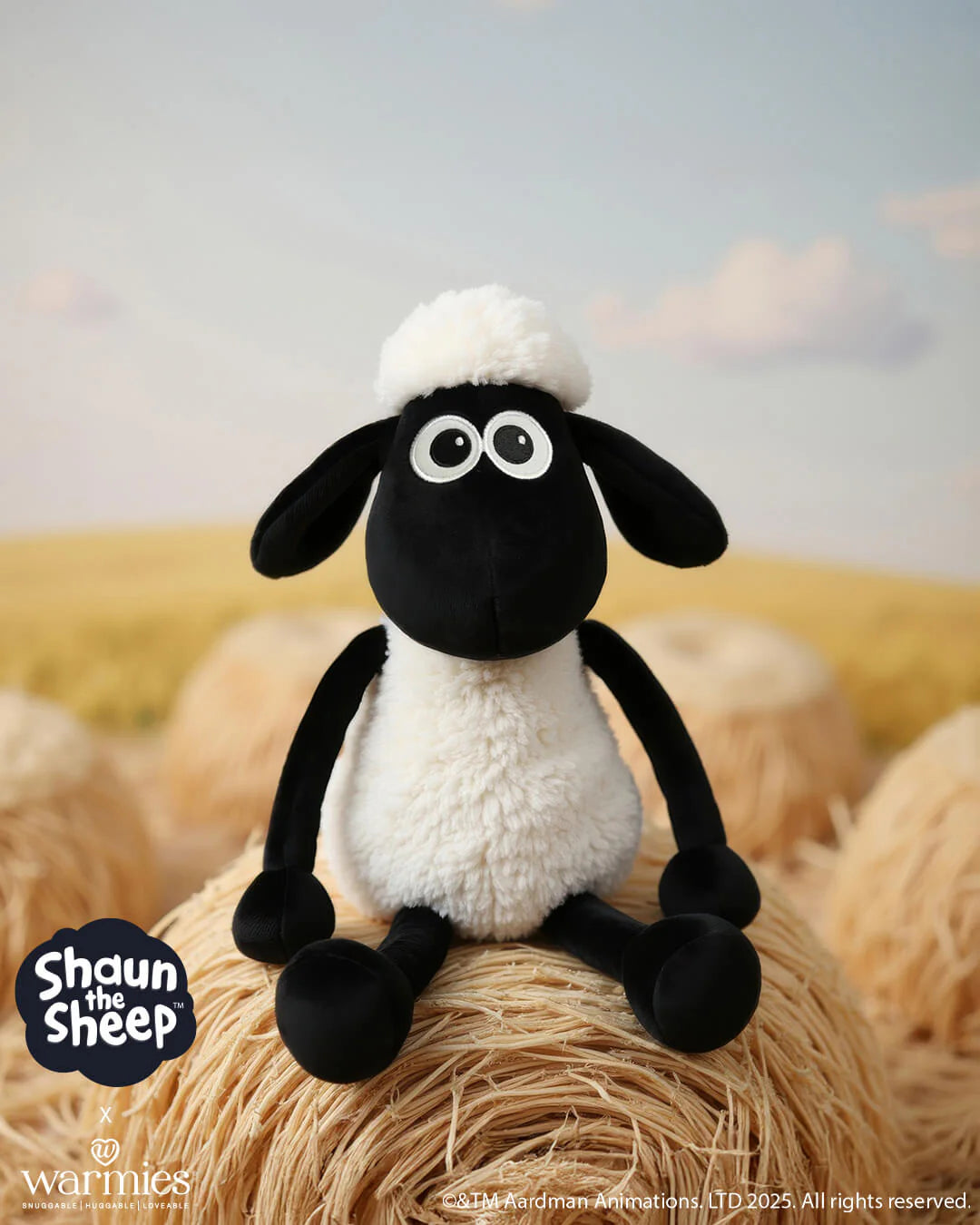 Plush toy sheep with black face and white wool sitting on a hay bale with a blurred field background.