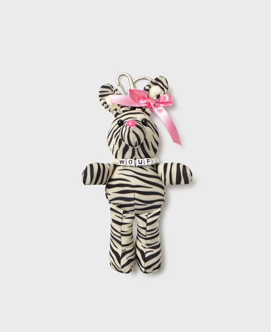 A zebra-print plush keychain shaped like a bear, featuring black and white stripes, a pink satin bow on its head, and a silver metal keyring at the top. The plush has a small white collar with the word 'MOUR' embroidered on it, set against a solid light gray background.