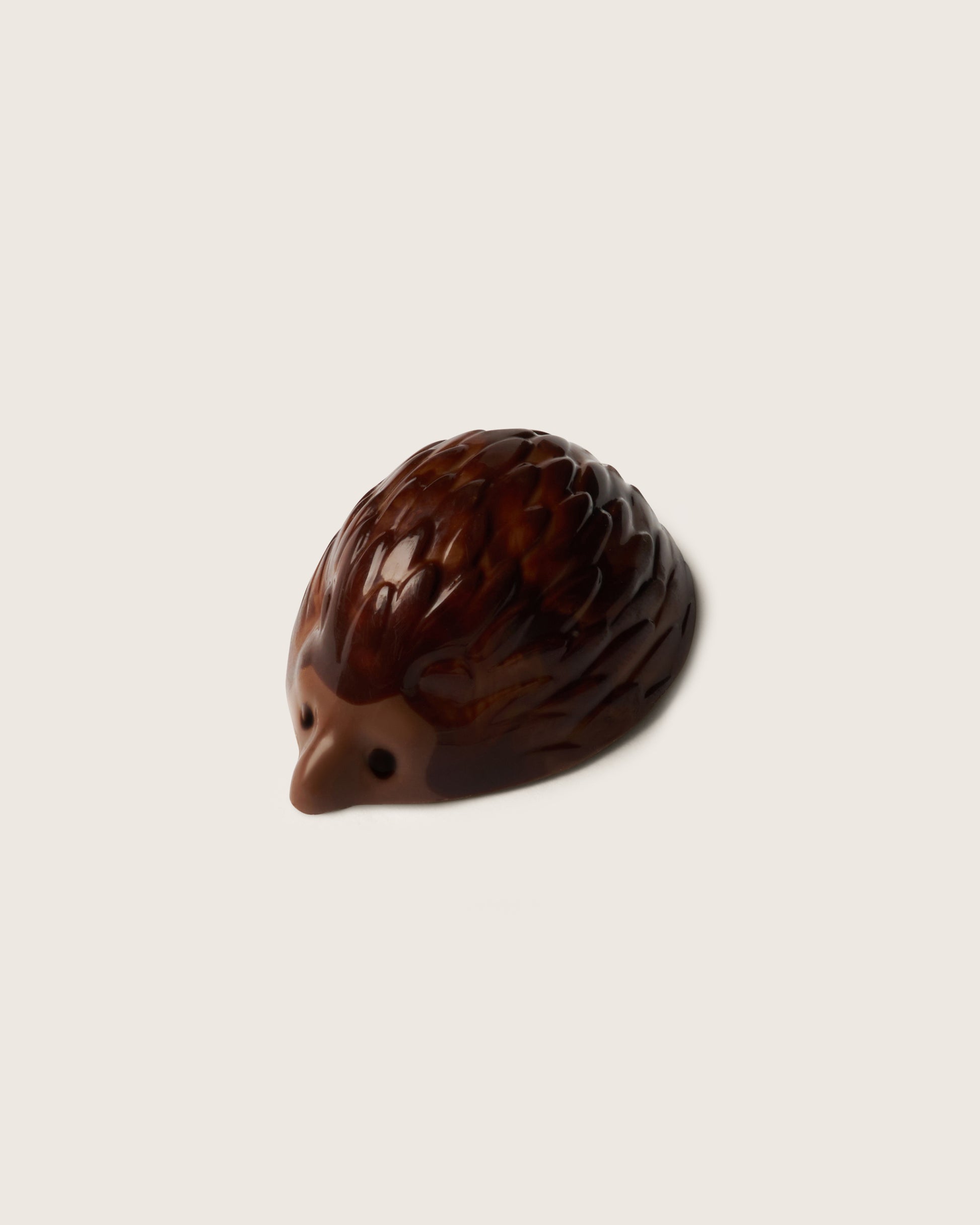 A small brown chocolate hedgehog praline with detailed spines and a smooth snout, photographed on a plain light beige background.