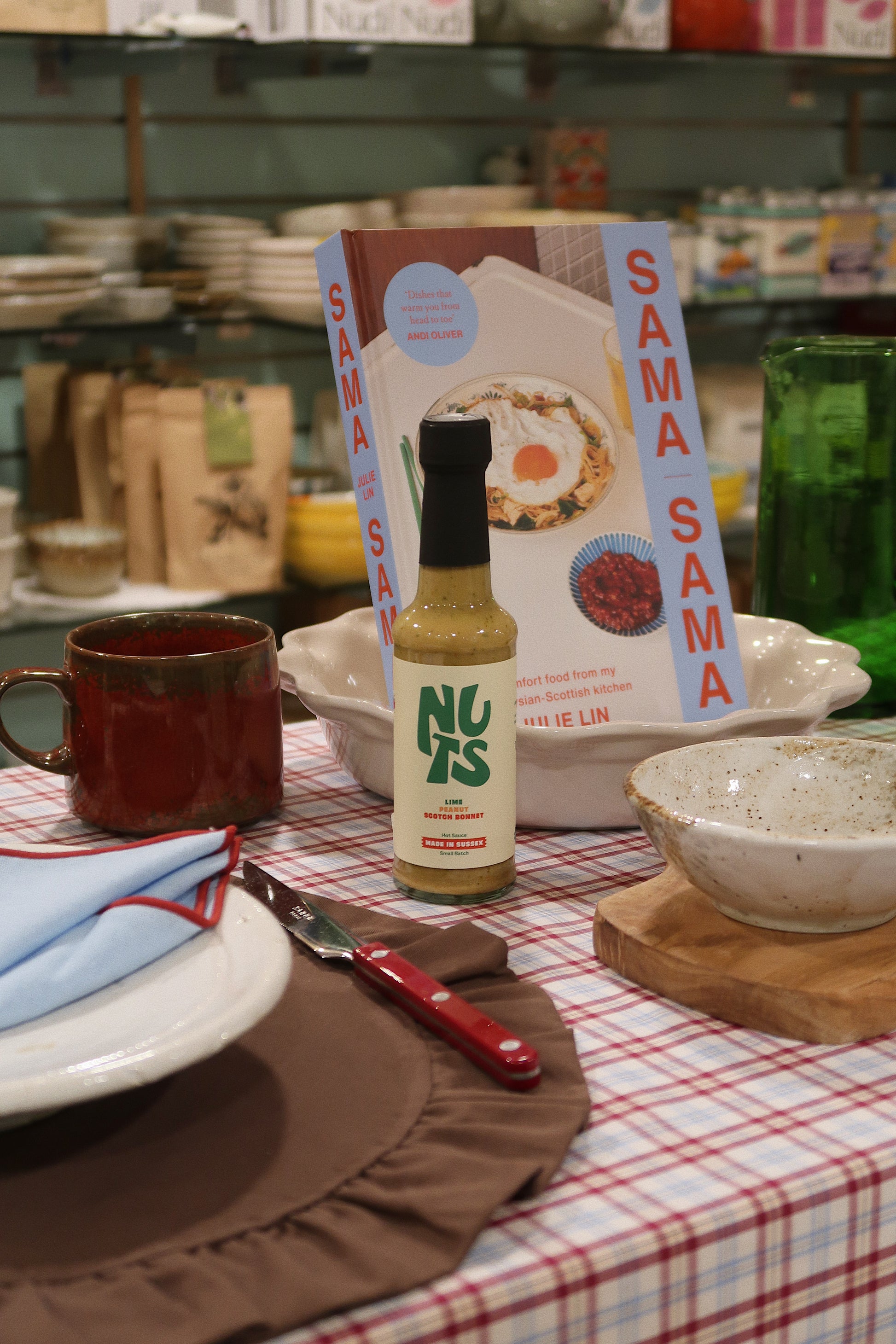 Table setting with 'Nuts' bottle, 'Sama Sama' book, and ceramic items in a store setting.