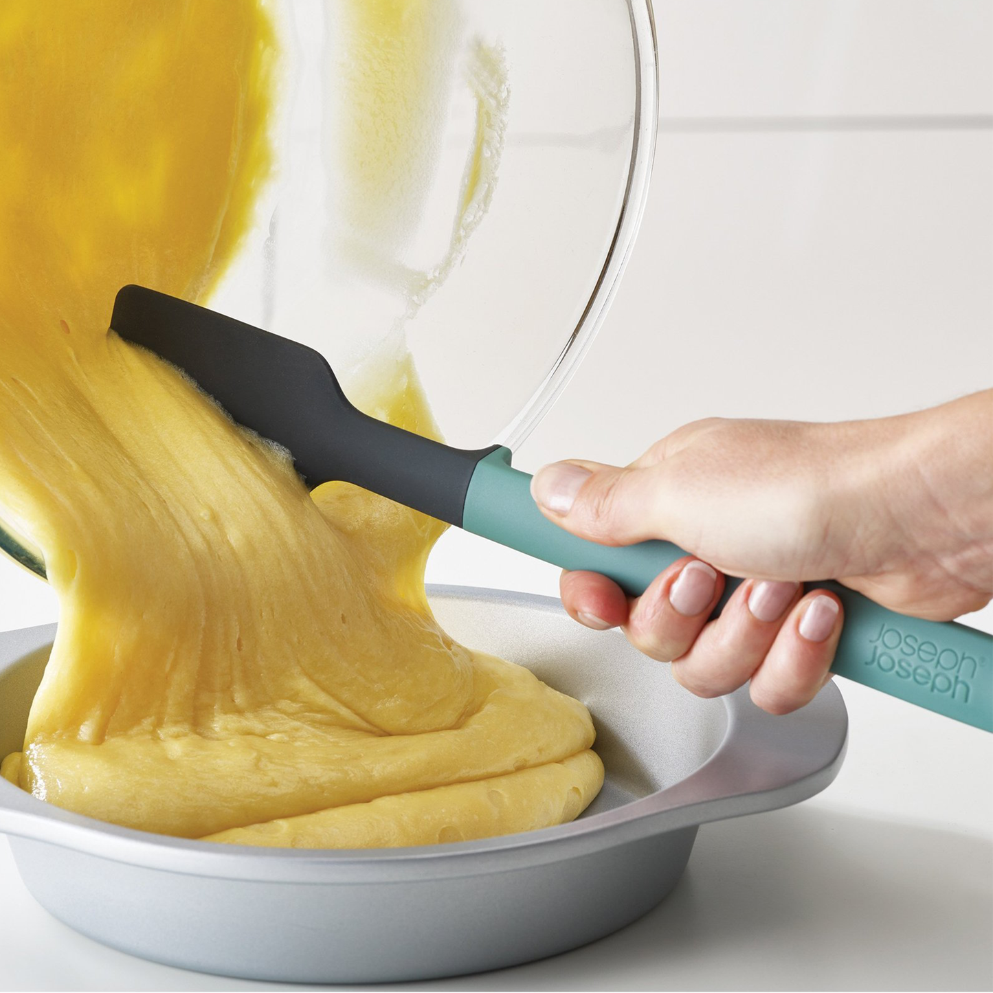 a person using the spatula to help pour cake batter from a glass mixing bowl into a cake pan