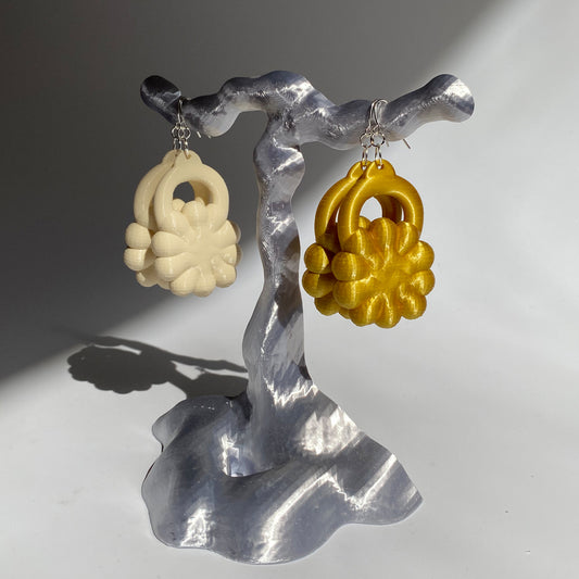 A pair of cream 3D printed earrings and a pair of gold digitally printed earrings on an earring stand photographed on plain background 